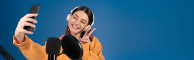 Teenager in headphones taking selfie on smartphone near studio microphone isolated on blue, banner 