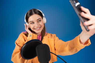 Teenager in headphones taking selfie on smartphone near blurred podcast microphone isolated on blue 