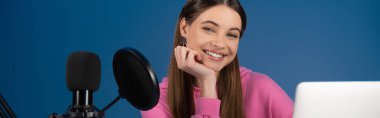 Teenager looking at camera near microphone and laptop isolated on blue, banner 
