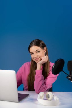 Positive teenager looking at camera near podcast microphone and laptop isolated on blue 