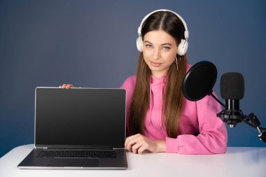 Teen podcaster in headphones sitting near laptop and microphone isolated on blue 