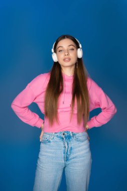 Teen girl in jeans and hoodie listening music in headphones isolated on blue 