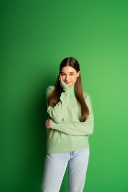 Smiling teenager in jeans and cozy jumper looking at camera on green background 