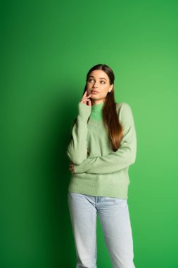Pensive teen girl in jumper and jeans looking away on green background 