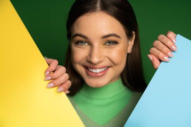 Pretty teen girl in jumper looking at camera and touching colorful paper isolated on green 