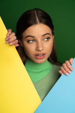 Pretty teen girl in warm jumper touching colorful paper isolated on green 