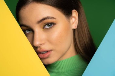 Portrait of brunette teen girl looking at camera near colorful paper isolated on green 