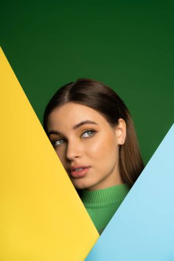 Brunette teenager in turtleneck posing near colorful paper isolated on green  clipart