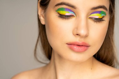 Teenage girl with colorful makeup and naked shoulder looking down isolated on grey 