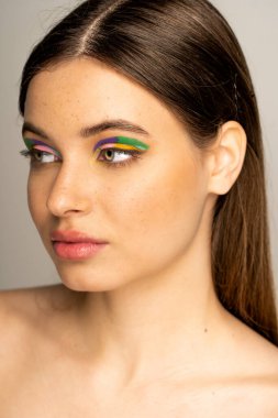 Brunette teen model with freckles and colorful eyeshadows looking away isolated on grey 
