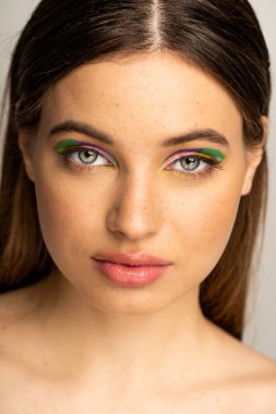Portrait of freckled teenager with multicolored makeup standing isolated on grey 