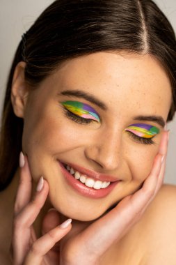 Pleased teen girl with colorful makeup touching face isolated on grey 