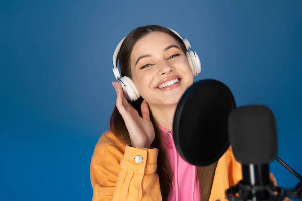 stock image Smiling teen girl in headphones looking at camera near blurred microphone isolated on blue 