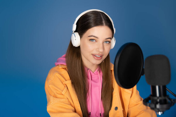 Smiling teen girl in headphones looking at camera near studio microphone isolated on blue 