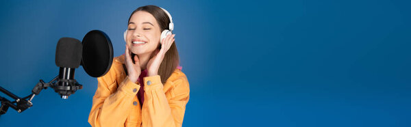 Joyful teenager in headphones standing near studio microphone isolated on blue, banner 