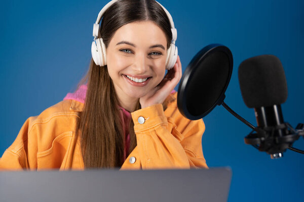 Улыбающийся подросток-подкастер в наушниках, смотрящий в камеру рядом с ноутбуком и микрофоном, изолированным на синем 