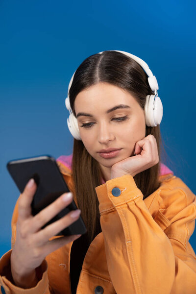 Teen girl in headphones using blurred smartphone isolated on blue 