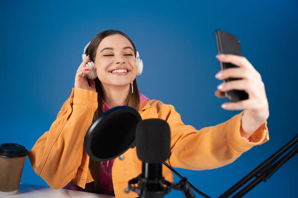Smiling teenager in headphones taking selfie on cellphone near studio microphone isolated on blue 
