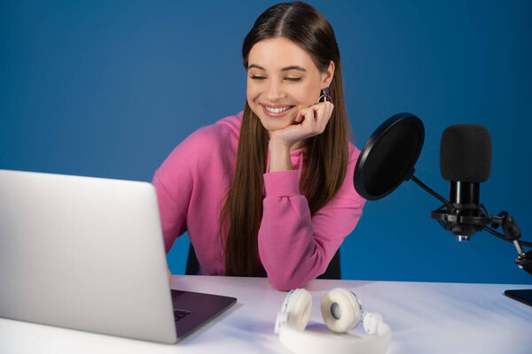 Smiling teenager using laptop near headphones and studio microphone isolated on blue 