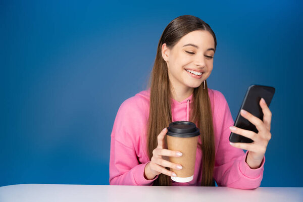 Smiling teenager holding takeaway coffee and using smartphone isolated on blue 