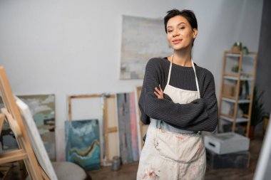 Artist in apron crossing arms and looking at camera in workshop 