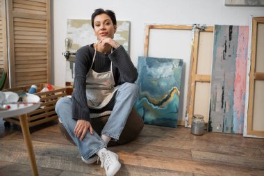 Tattooed artist in apron sitting on floor near paintings in studio 