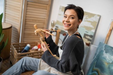 Cheerful artist holding wooden doll and looking at camera near drawings in workshop 
