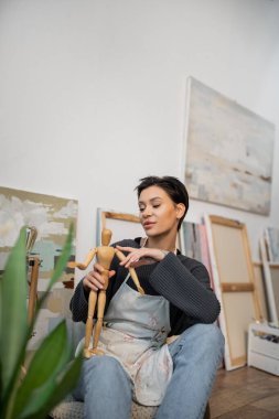 Tattooed artist in apron holding wooden doll near plant and paintings in workshop 