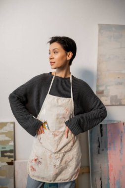 Young artist in apron with paintbrushes standing in workshop 