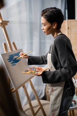 Side view of brunette artist painting on canvas in studio 