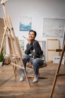 Positive artist holding paintbrush and looking at camera in workshop  clipart