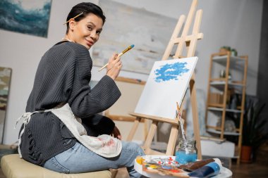 Pleased artist holding paintbrush and looking at camera near easel on canvas 