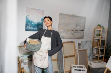 Short haired artist in apron holding basket with equipment near drawings in studio 
