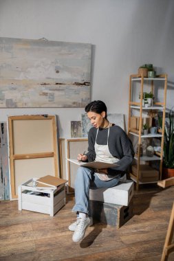 Brunette artist in apron drawing on sketchbook in workshop 