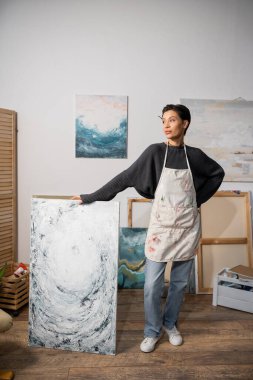 Short haired artist standing with hand on hip near painting in workshop 
