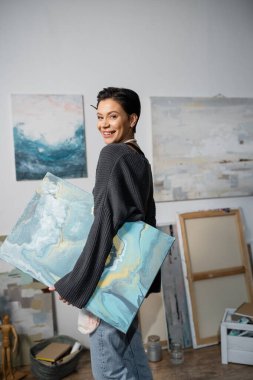 Happy artist looking at camera and holding painting in studio  clipart