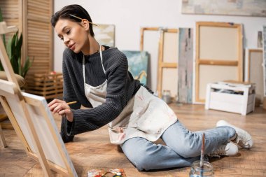 Brunette artist in dirty apron painting on canvas while sitting on floor 