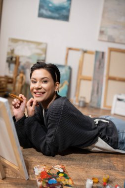 Cheerful artist with paintbrushes smiling at camera near paints and canvas on floor in studio 
