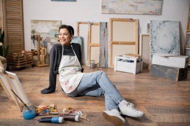 Positive artist in apron sitting on floor near paints and painting in workshop 