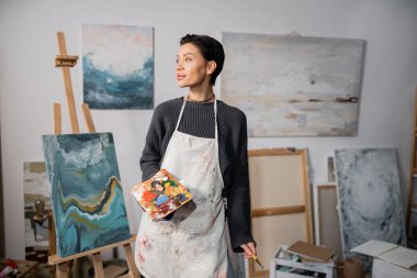 Young artist holding palette and paintbrush near painting on easel 