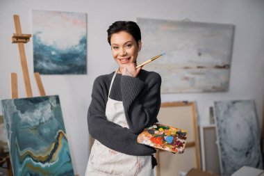 Cheerful tattooed artist holding paintbrush and palette in studio 