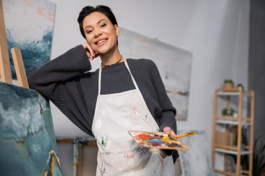 Smiling artist in dirty apron holding paintbrush and palette near painting in studio 
