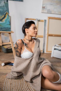 Sexy short haired artist in sweater and bra sitting on floor in workshop 
