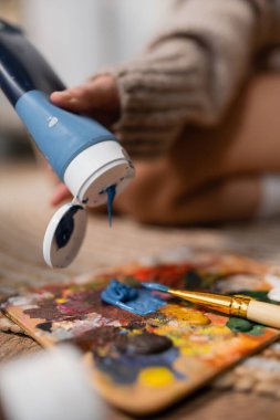 Cropped view of artist pouring paint on palette near paintbrush in workshop 