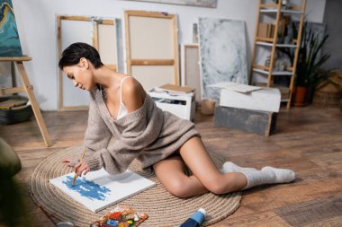 Side view of sexy artist in sweater painting on floor in workshop 