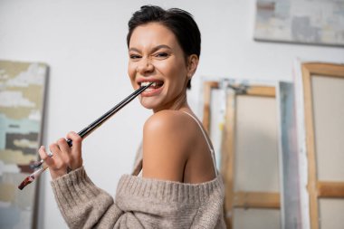 Cheerful artist in sweater and bra holding paintbrushes in studio  clipart