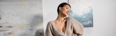 Sexy tattooed artist in sweater standing near paintings in studio, banner 