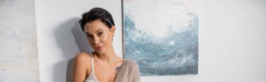 Sexy artist with pencil looking at camera near paintings in studio, banner 