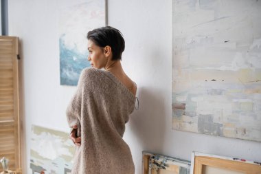 Side view of sexy brunette artist in sweater standing near paintings in studio 
