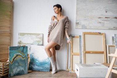 Sensual artist in sweater and paint on hand standing near paintings in workshop 